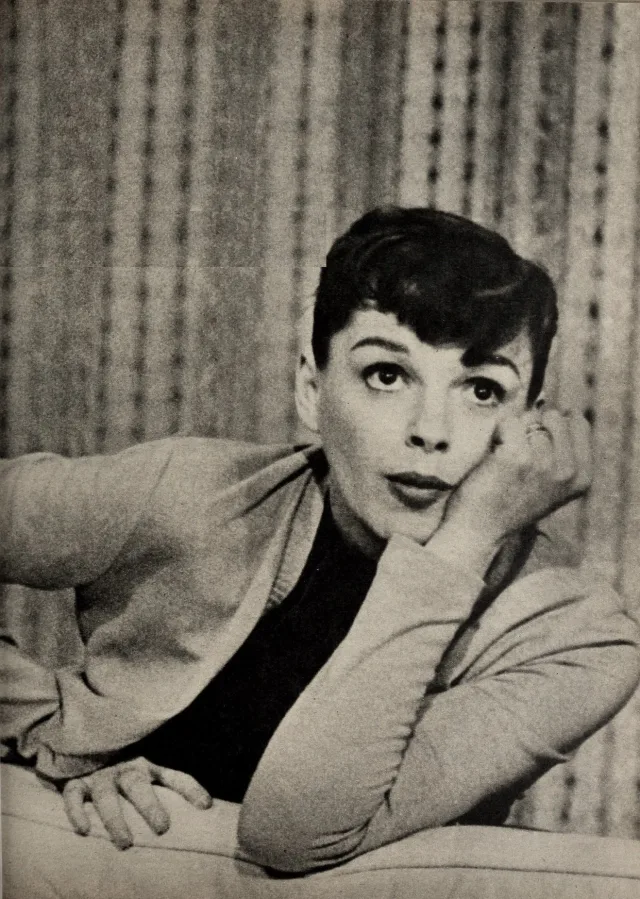Black and white portrait of a short-haired woman resting her chin on one hand and looking upward against a striped curtain.