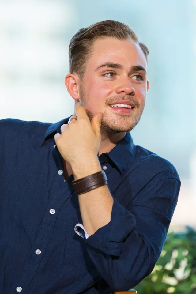 A smiling young man with short swept-back hair and light stubble, in a navy shirt, glancing to one side with a hand raised near his chin.