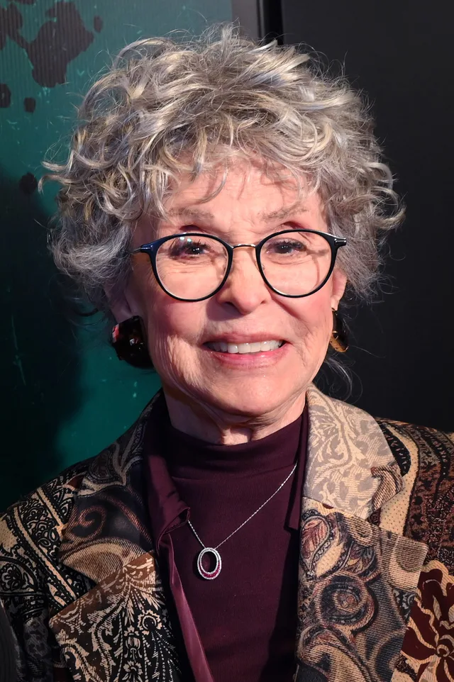 Smiling older woman with short curly grey hair and round dark-framed glasses, wearing a patterned jacket over a plum polo neck and a pendant necklace.