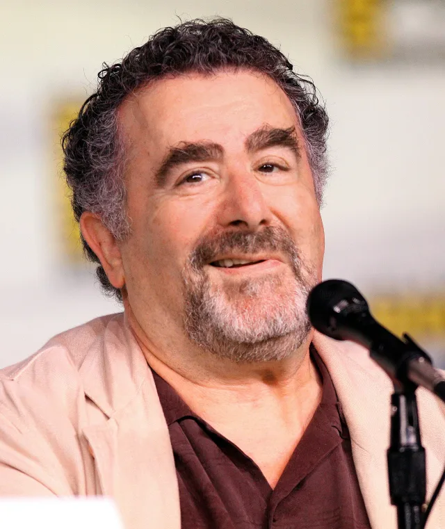 Smiling man with curly grey-black hair and a goatee, in a beige jacket, speaking at a microphone on a convention panel