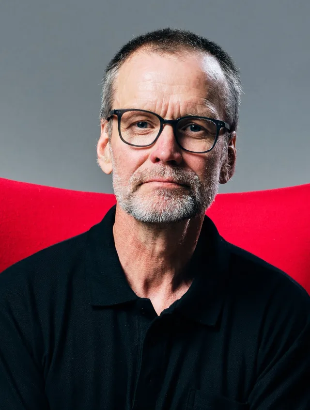Older man with short grey hair, a grey beard and dark-rimmed glasses, in a black polo shirt in front of a red chair