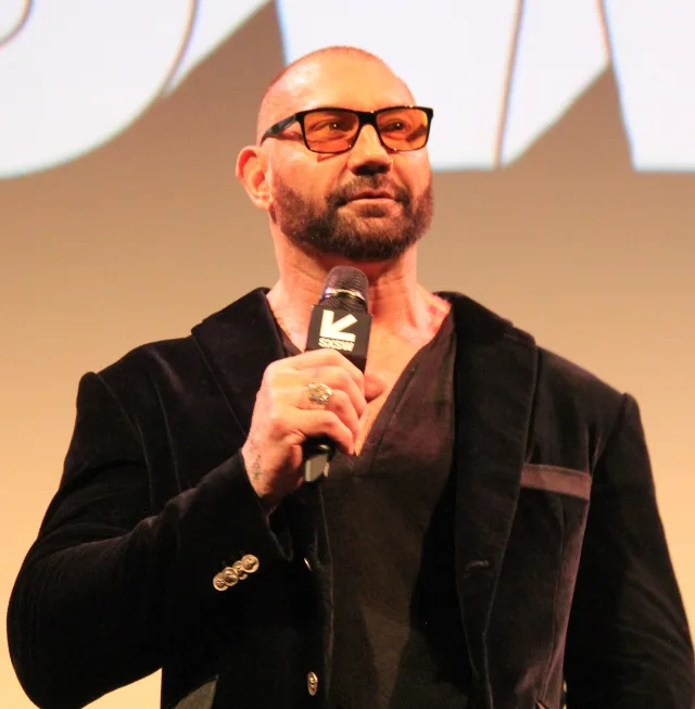 A bald, bearded man in tinted glasses and a dark velvet jacket holds a microphone while speaking on a stage.
