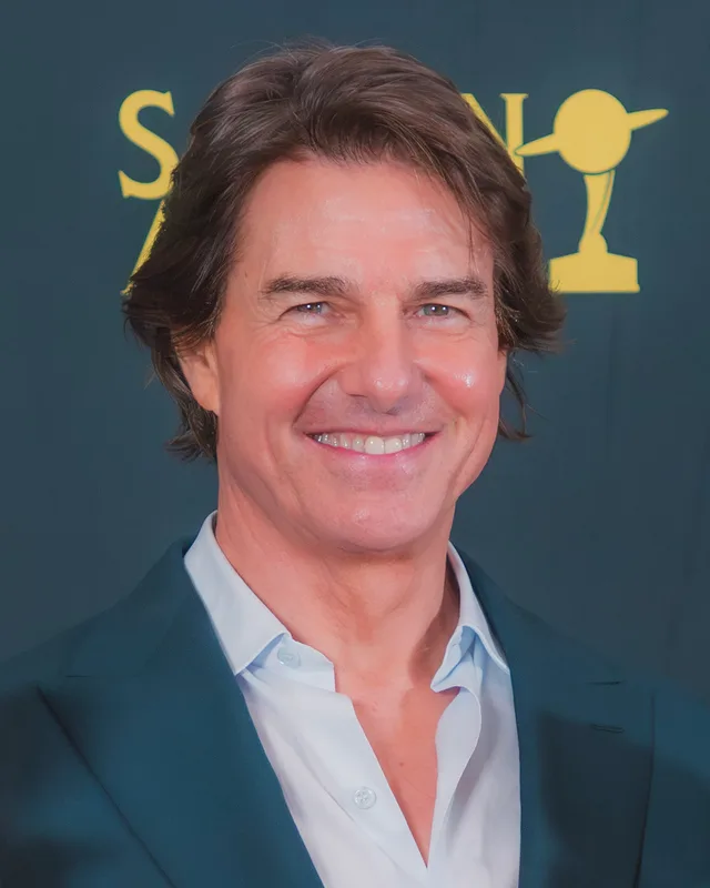 Head and shoulders photograph of Tom Cruise smiling, with swept-back brown hair, in a teal jacket and open-collared white shirt against a dark backdrop.