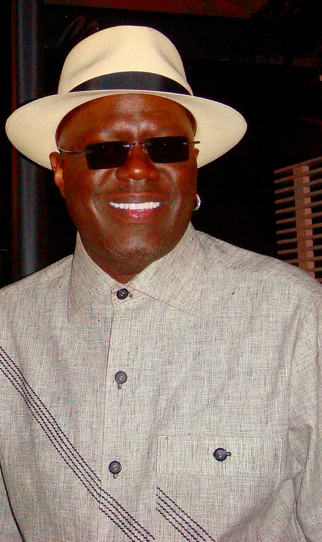 A smiling man in a cream fedora with a dark band, dark rimless sunglasses and a grey buttoned shirt with dark stitching.