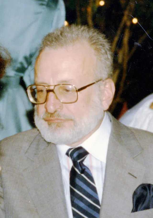 Candid colour photograph of an older bearded man with glasses, looking down, in a grey jacket and striped tie.