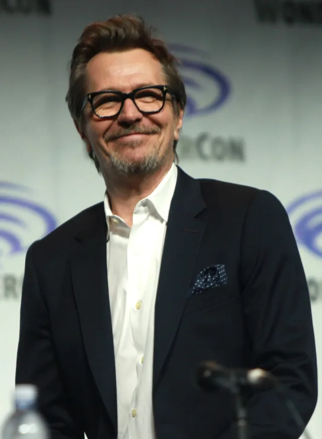 Gary Oldman smiling in black-framed glasses, a navy jacket and white open-necked shirt, seated at a convention panel.