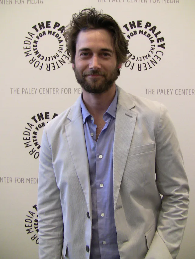 Ryan Eggold standing with tousled hair and a beard, in a light grey blazer over a blue shirt, in front of a white event wall.