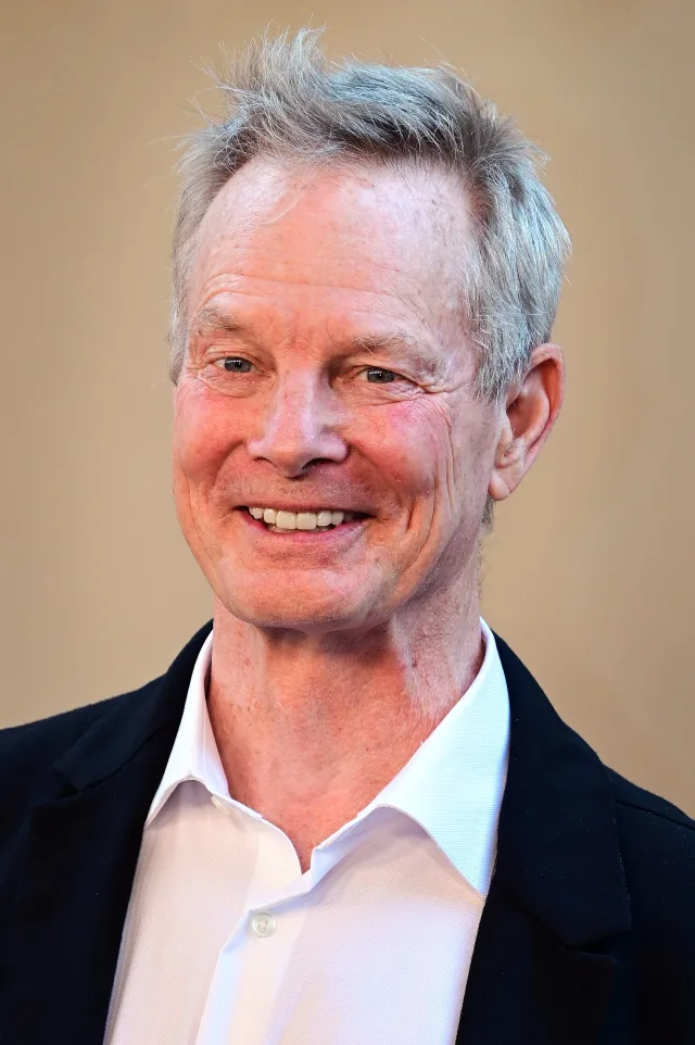 Close-up portrait of a smiling older man with swept grey hair, wearing a white open-collared shirt and dark jacket against a warm beige background.