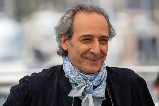 A smiling man with swept-back grey hair, wearing a dark jacket and a patterned blue and white scarf, photographed outdoors with a blurred background.