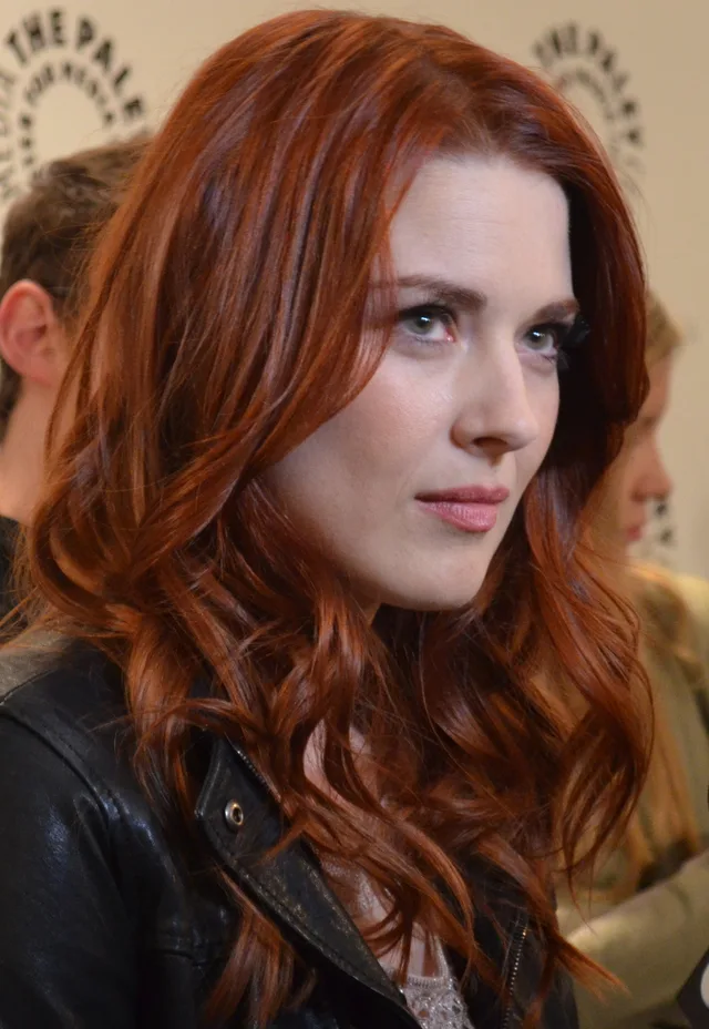 Woman with long wavy auburn hair and a black leather jacket, looking to one side at a press event.