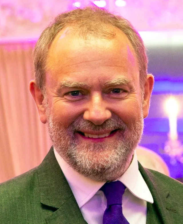 Close portrait of a smiling man with a grey beard and receding hair, wearing a green jacket and purple tie against a softly lit pink background.
