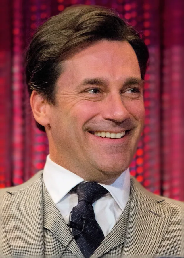 Close portrait of a smiling man with swept-back dark hair, wearing a houndstooth waistcoat and tie, in front of a red curtain.