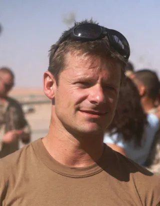 Close portrait of a man with short light-brown hair and sunglasses pushed up on his head, wearing a tan t-shirt outdoors in bright sunshine.