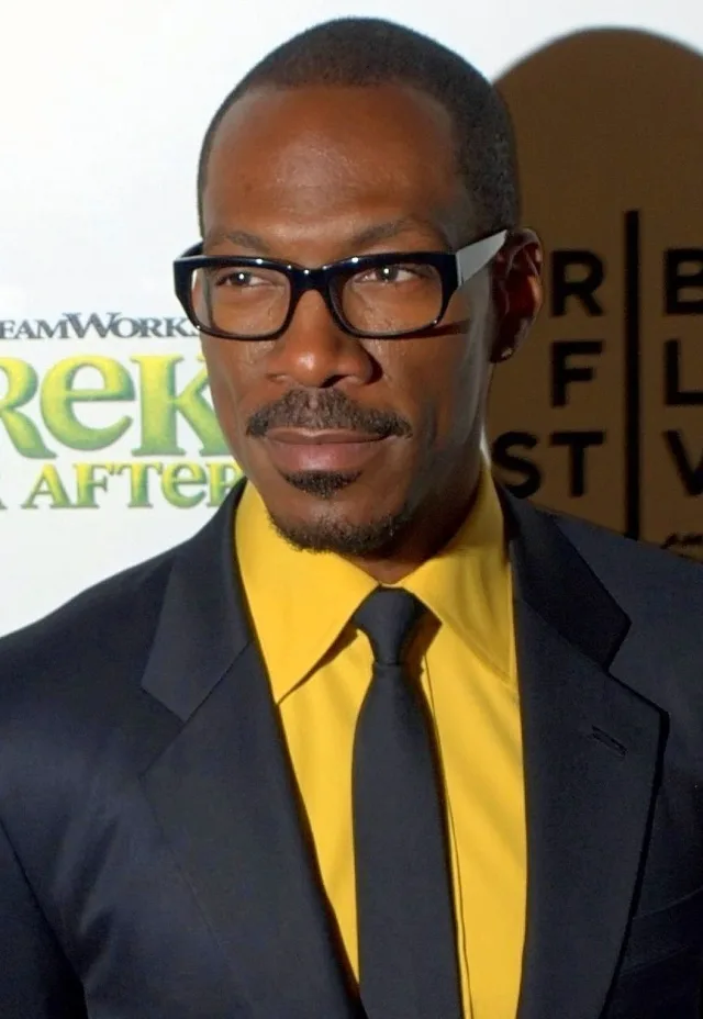 Eddie Murphy in black-framed glasses, a dark suit and a yellow shirt with a black tie, at a press event.