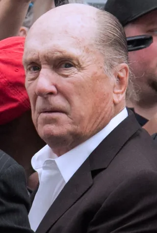 Close photograph of an elderly bald man with a serious expression, wearing a white open-collared shirt and dark jacket, with a crowd behind him.