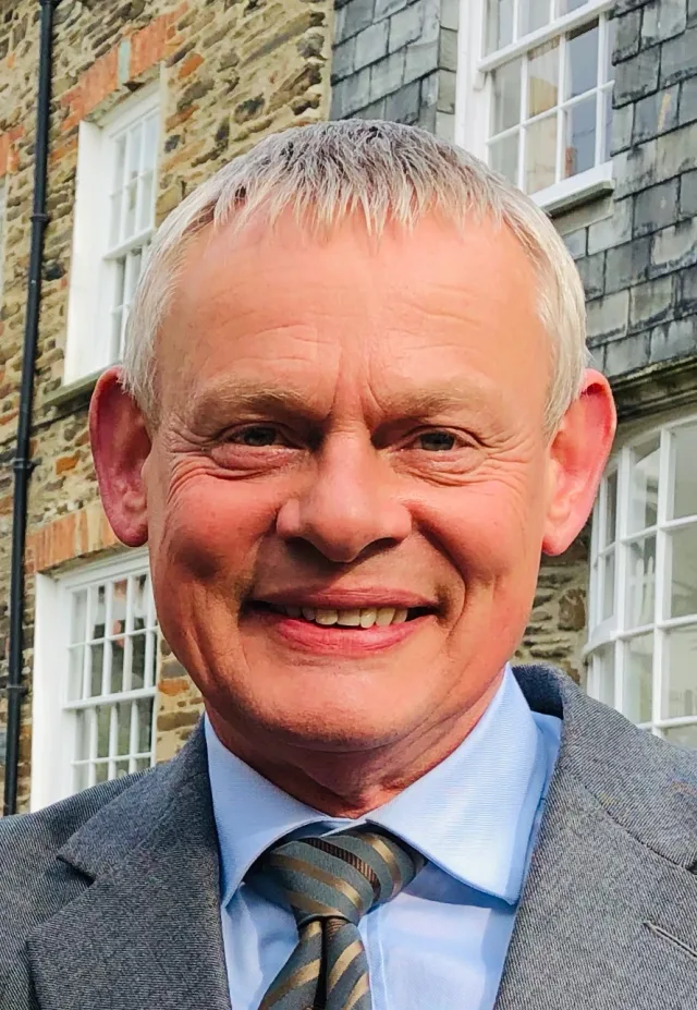 Close portrait of a smiling grey-haired man in a grey jacket, blue shirt and striped tie, in front of a stone building.