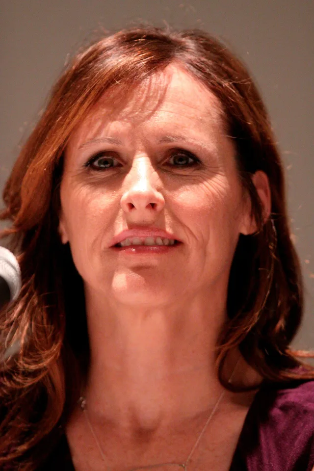 Close photograph of a woman with shoulder-length auburn hair and a purple top, seated near a microphone against a plain grey background.