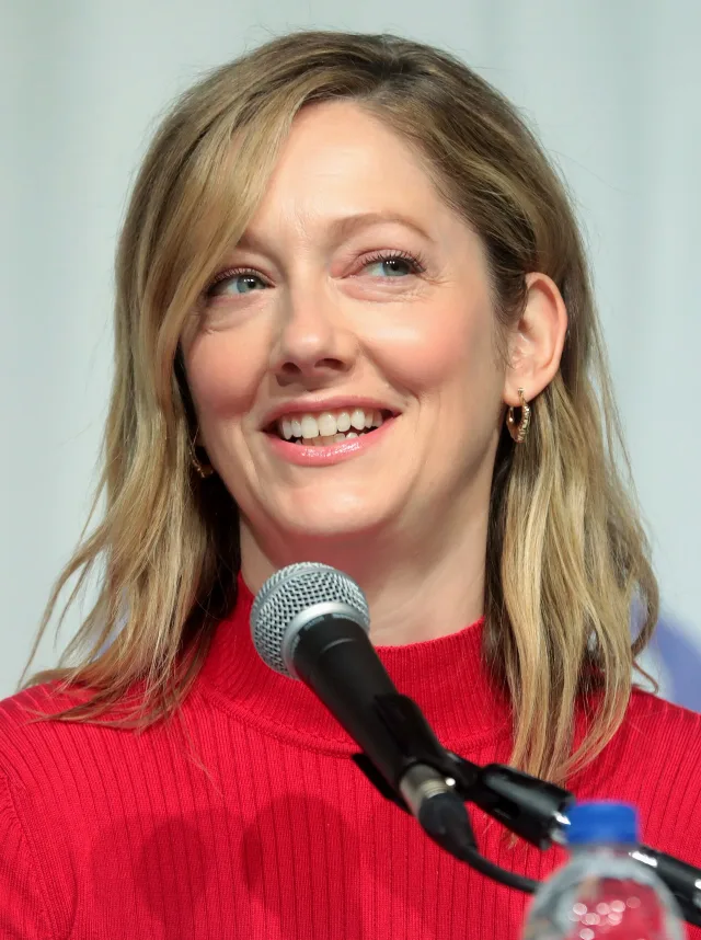 Judy Greer smiling at a panel microphone, shoulder-length fair hair, wearing a red ribbed top.