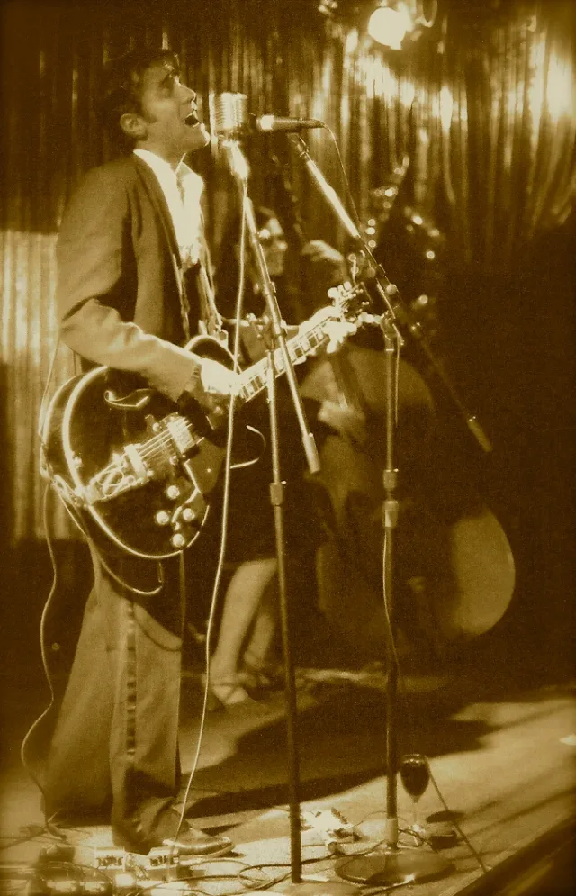 Sepia concert photograph of a man singing into a microphone while playing a dark electric guitar on a stage with a curtain behind him.