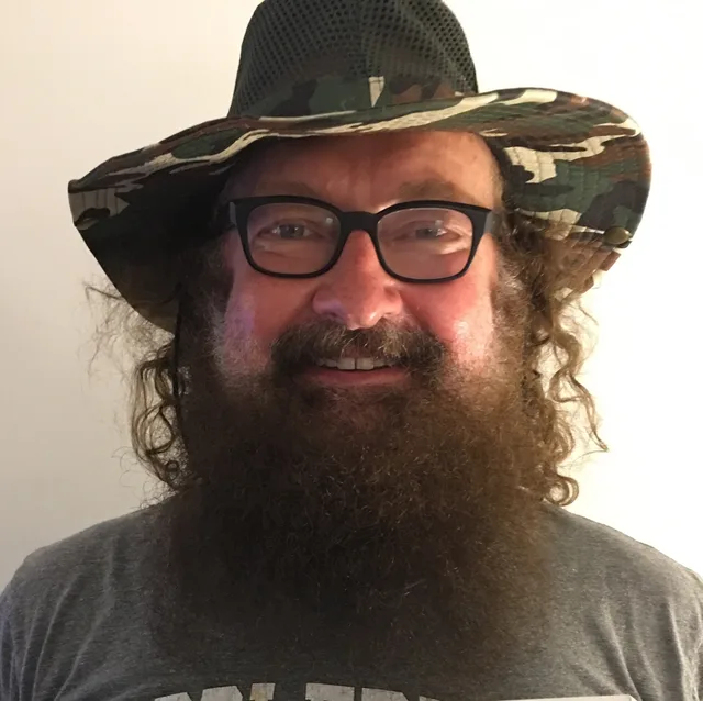 Head and shoulders photograph of a smiling man with a long brown beard, dark-framed glasses and a camouflage bucket hat, wearing a grey T-shirt.