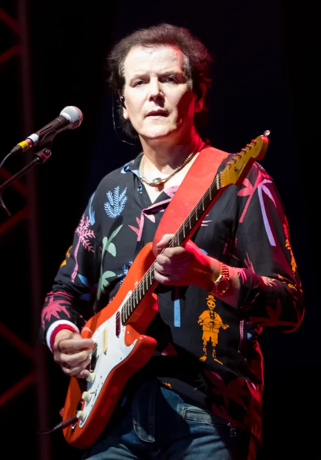 Trevor Rabin on stage playing a red and white electric guitar, in a patterned dark shirt, beside a microphone stand.