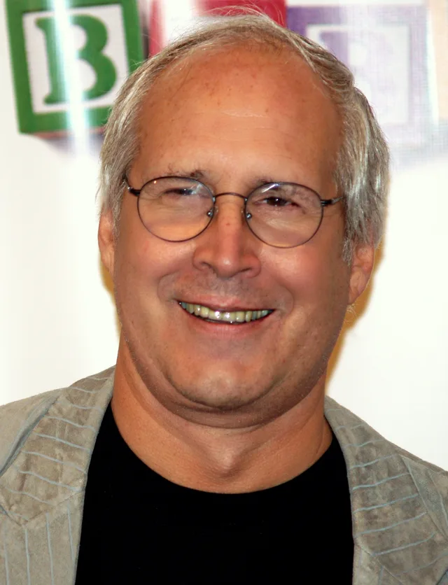 Chevy Chase smiling in round wire glasses and a grey-green jacket over a black top, with coloured alphabet blocks blurred behind him.