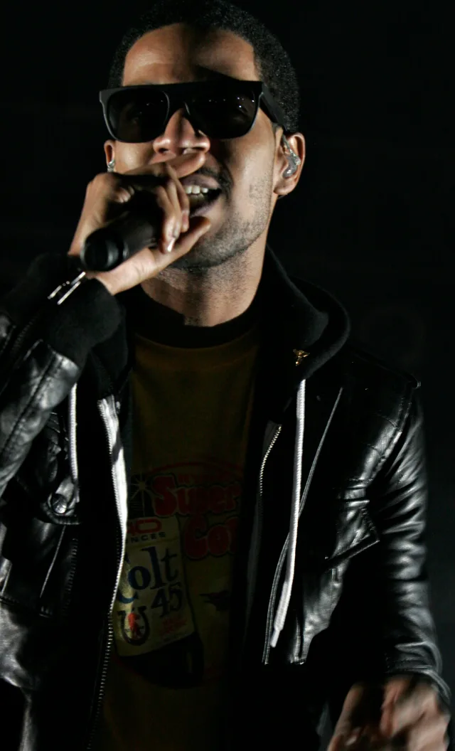 Kid Cudi performing on stage in sunglasses and a black leather jacket, singing into a handheld microphone against a dark background.