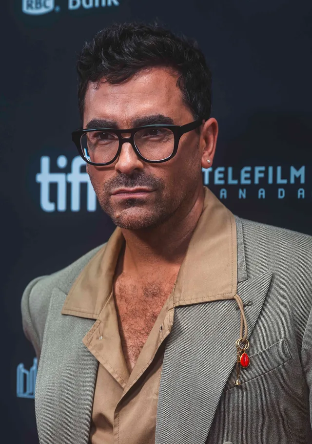 A man with short dark hair, stubble and black-framed glasses, wearing a sage jacket over a tan shirt, standing in front of a festival backdrop.