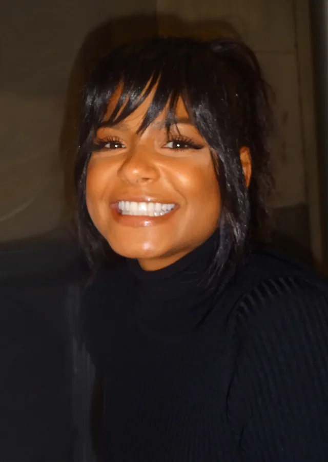 Smiling woman with dark, choppy shoulder-length hair and a fringe, wearing a black ribbed polo-neck jumper, photographed close up indoors.