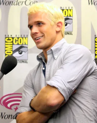 A smiling man with short bleached blond hair and a grey shirt with rolled sleeves, arms crossed, speaking beside a microphone at a convention press wall