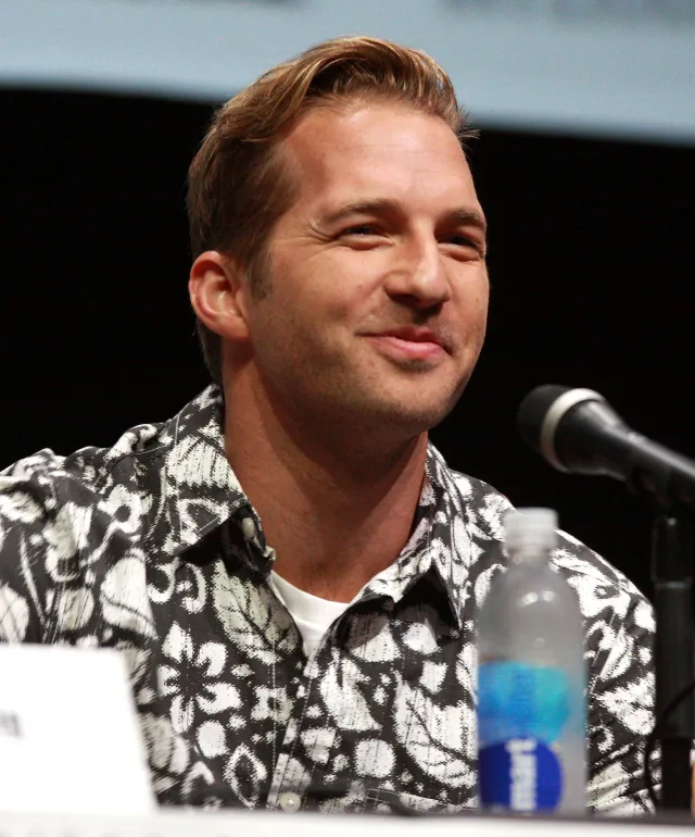 Smiling man with swept-back fair hair in a black and white floral shirt, seated at a panel microphone.
