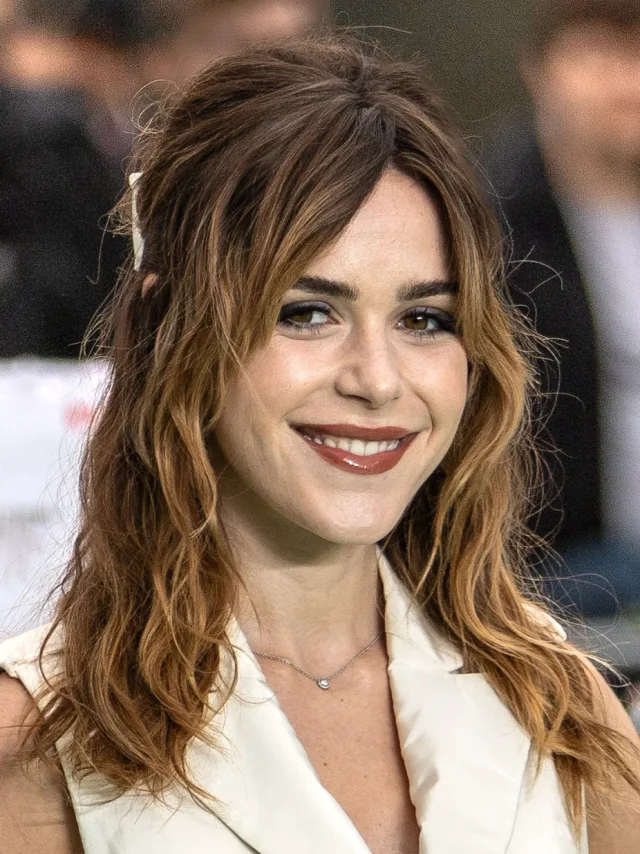 Smiling woman with long wavy brown hair, dark lipstick and a cream sleeveless collar, with a delicate silver pendant necklace.
