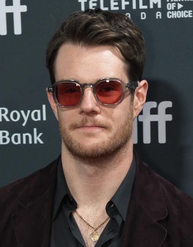 Connor Swindells with short dark hair and a light beard, wearing red-tinted glasses and a dark jacket in front of a festival backdrop.