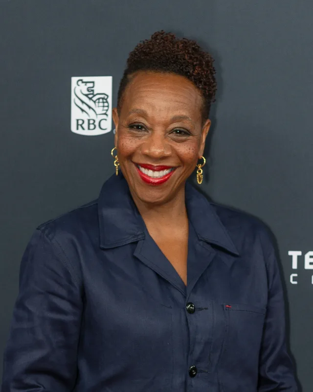 Smiling woman with short curly hair, red lipstick and gold hoop earrings, wearing a navy button-up jacket, in front of a dark event backdrop.