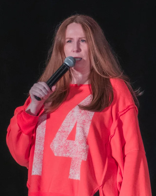 Woman with long red-brown hair in a coral sweatshirt speaking into a microphone against a dark backdrop.