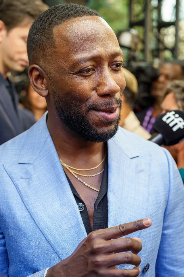 Close portrait of a bearded man in a light blue blazer and gold chains, speaking to reporters at a festival.