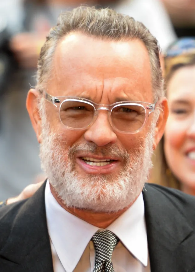 Close portrait of a man with swept-back grey hair, a white beard and clear-framed glasses, in a dark suit and patterned tie, smiling.