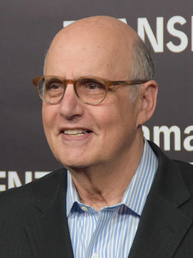 Close photograph of a smiling bald man in tortoiseshell glasses, a dark jacket and a blue striped shirt, standing in front of a press backdrop.