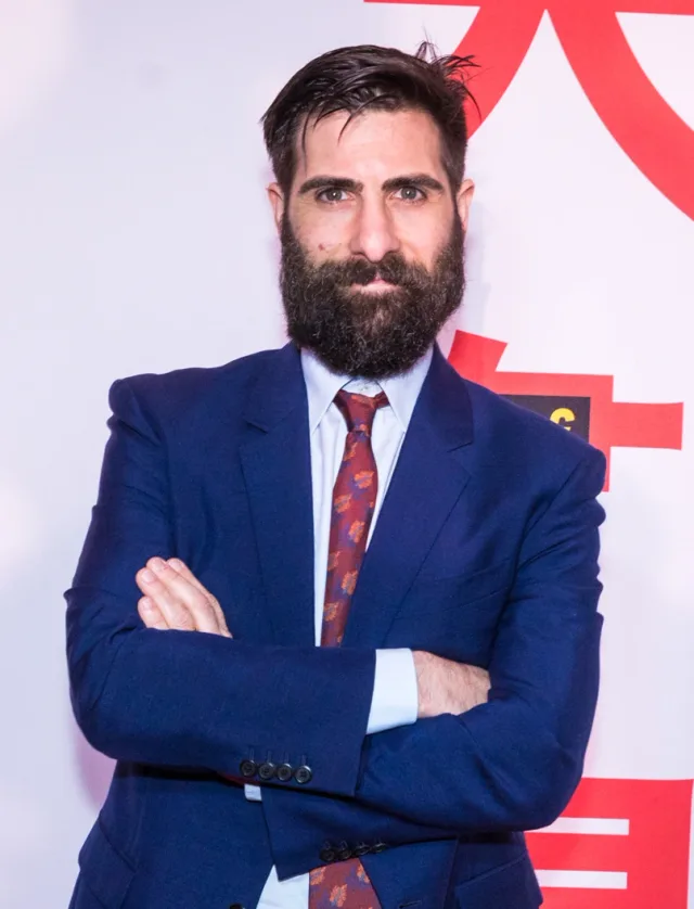 Bearded man with swept dark hair in a navy suit, pale blue shirt and patterned tie, arms folded, standing in front of a white and red backdrop.