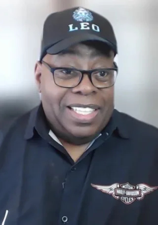 A smiling man in dark-framed glasses and a black cap and navy polo shirt, looking at the camera in a video-call style frame.