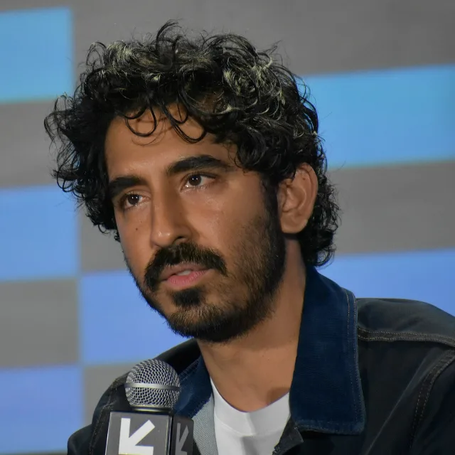 Close portrait of Dev Patel at a press panel, dark curly hair and a beard, a microphone in front of him.