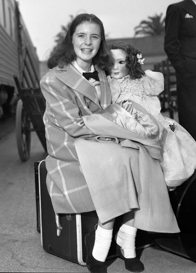 Black and white photograph of a young girl smiling, sitting on a suitcase outdoors in a checked coat with a large doll on her lap.