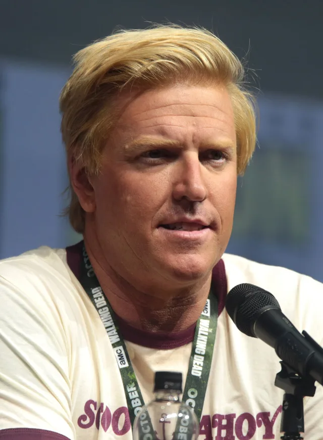 A blond man in a cream and maroon ringer T-shirt with a convention lanyard, speaking at a panel table beside a microphone.