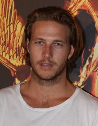 Head and shoulders photograph of a man with swept-back light brown hair and stubble, wearing a white t-shirt, in front of an orange and gold backdrop.