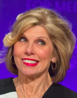 Close portrait of a smiling woman with a layered blonde bob, bright red lipstick and gold hoop earrings against a purple background.
