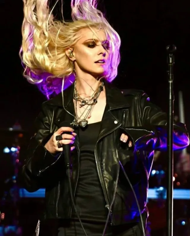 A blonde woman performing on stage in a black leather jacket, holding a microphone, her hair flung upward and lit in purple.
