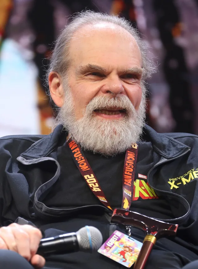 An older man with a long white beard and grey hair tied back, smiling as he speaks into a microphone at a convention panel.