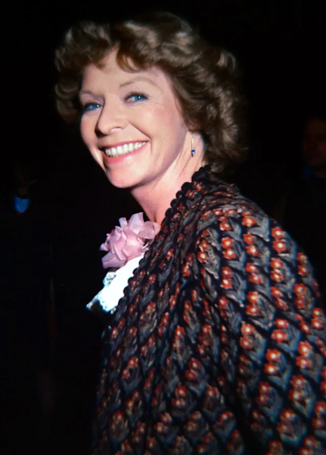 Smiling woman with short wavy brown hair and blue eyes, glancing back over her shoulder, wearing a patterned dark jacket with a pink bow at the neck.