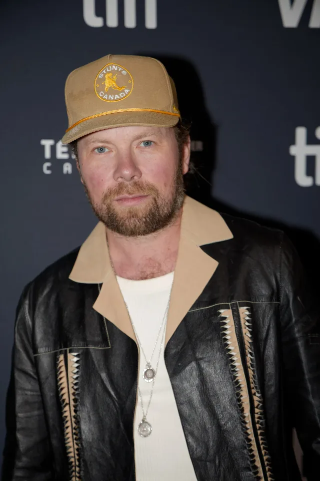A bearded man with light eyes in a tan cap and a black leather jacket with tan trim, standing in front of a festival backdrop
