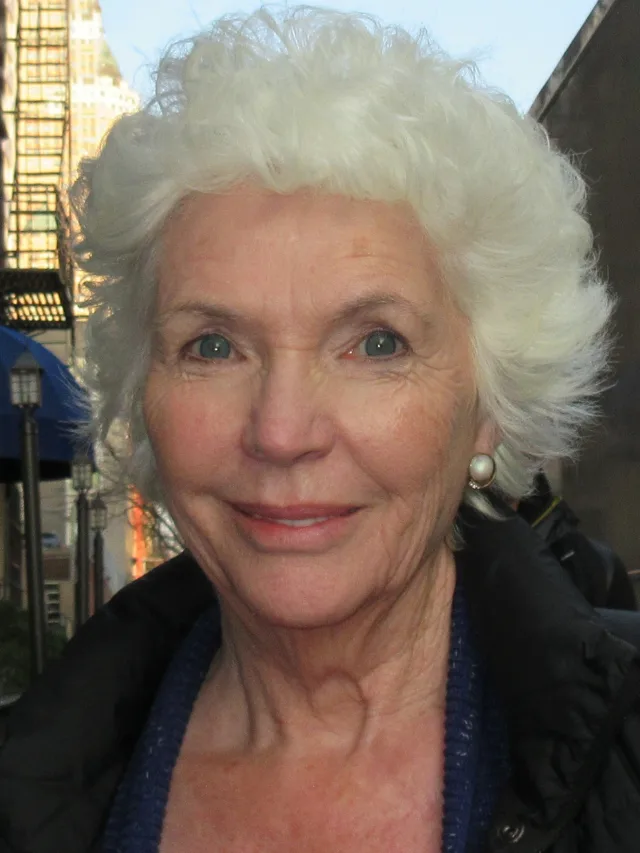 Close portrait of Fionnula Flanagan, smiling, with short white hair, pearl earrings and a dark jacket on a city street.
